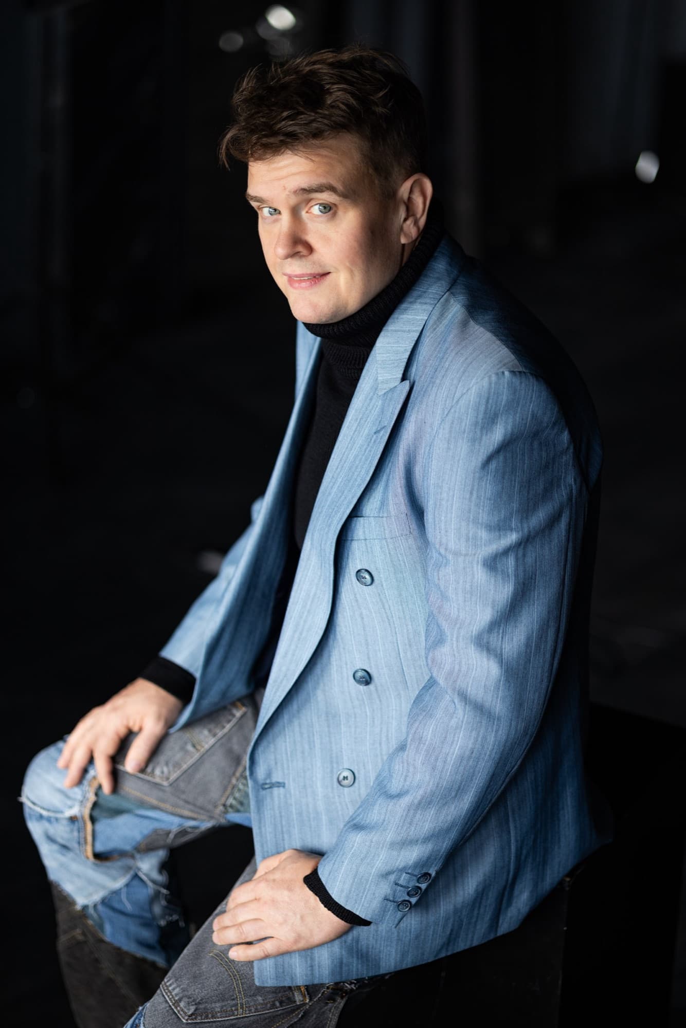 Mies vaaleansinisessä bleiserissä ja mustassa poolopaidassa istuu sivuttain tummaa taustaa vasten ja katsoo olkansa yli kameraan.
