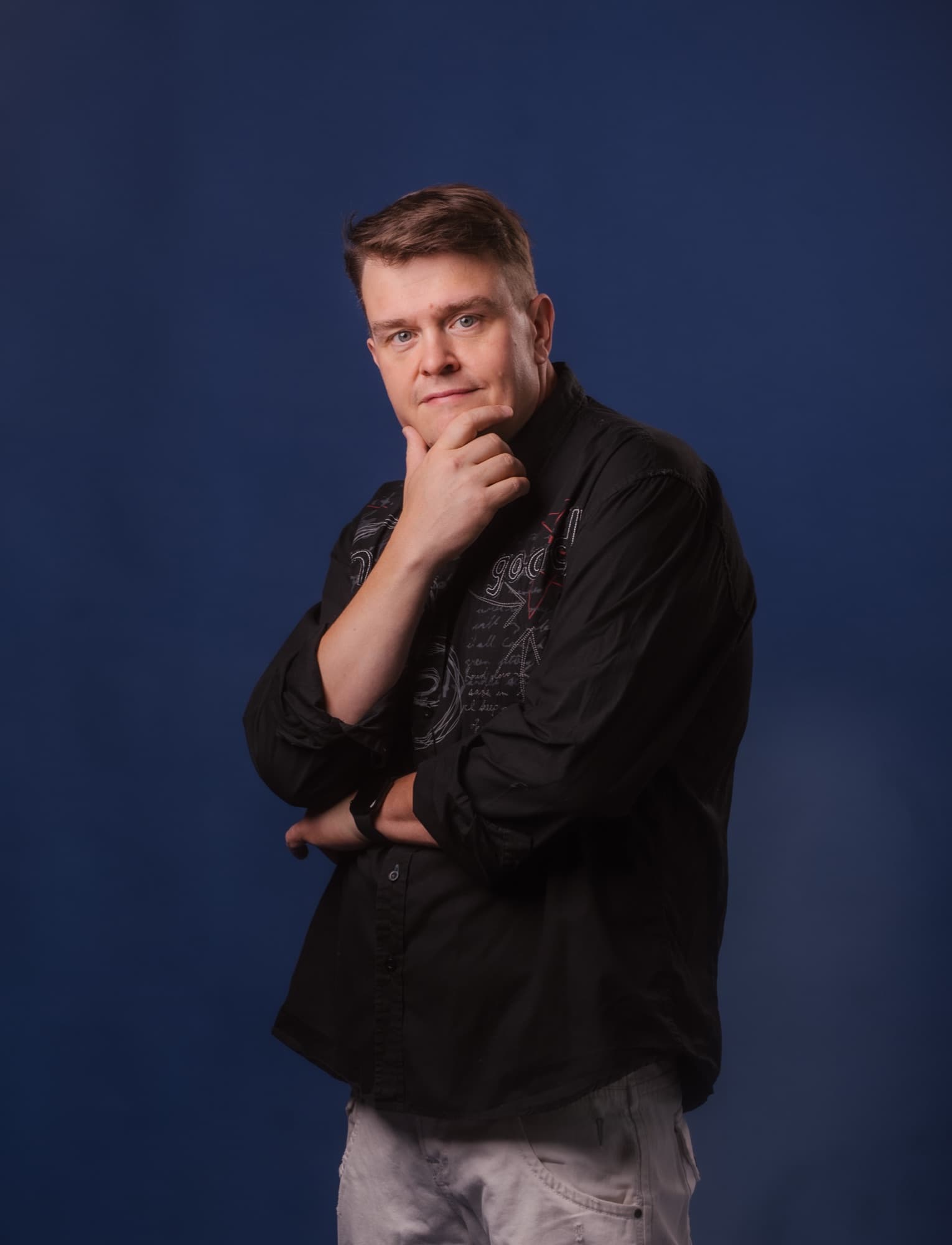 Studiokuva miehestä tummansinistä taustaa vasten, seisoo sivuttain ja koskettaa leukaansa mietteliäässä asennossa.